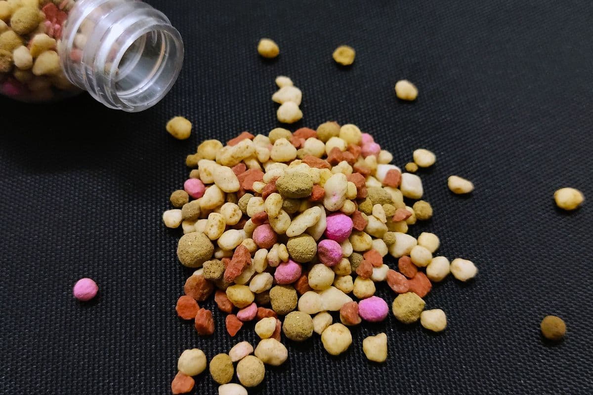 Grânulos de fertilizante de várias cores, em tons de branco, bege, marrom, laranja e rosa, espalhados sobre um tecido preto ao lado de um frasco tombado.