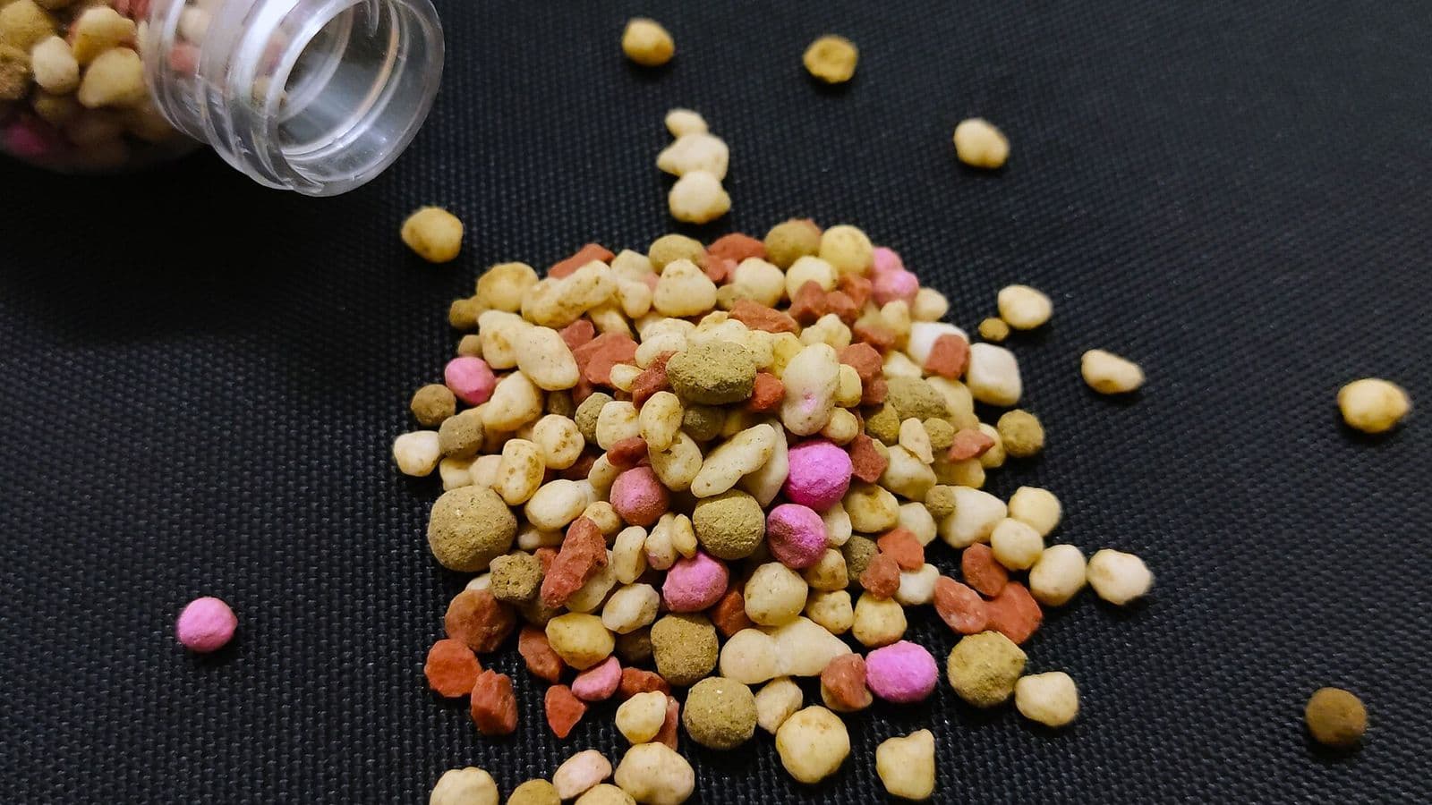 Grânulos de fertilizante de várias cores, em tons de branco, bege, marrom, laranja e rosa, espalhados sobre um tecido preto ao lado de um frasco tombado.