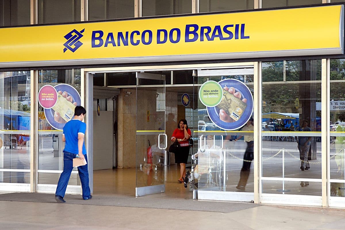 Fachada de agência do Banco do Brasil com letreiro amarelo e portas de vidro, com um homem entrando e uma mulher saindo da agência