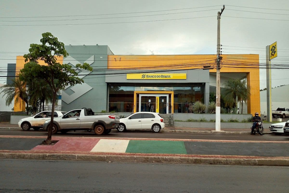 Fachada amarela e cinza de agência do Banco do Brasil, com carros estacionados em frente, poste com fios e céu nublado