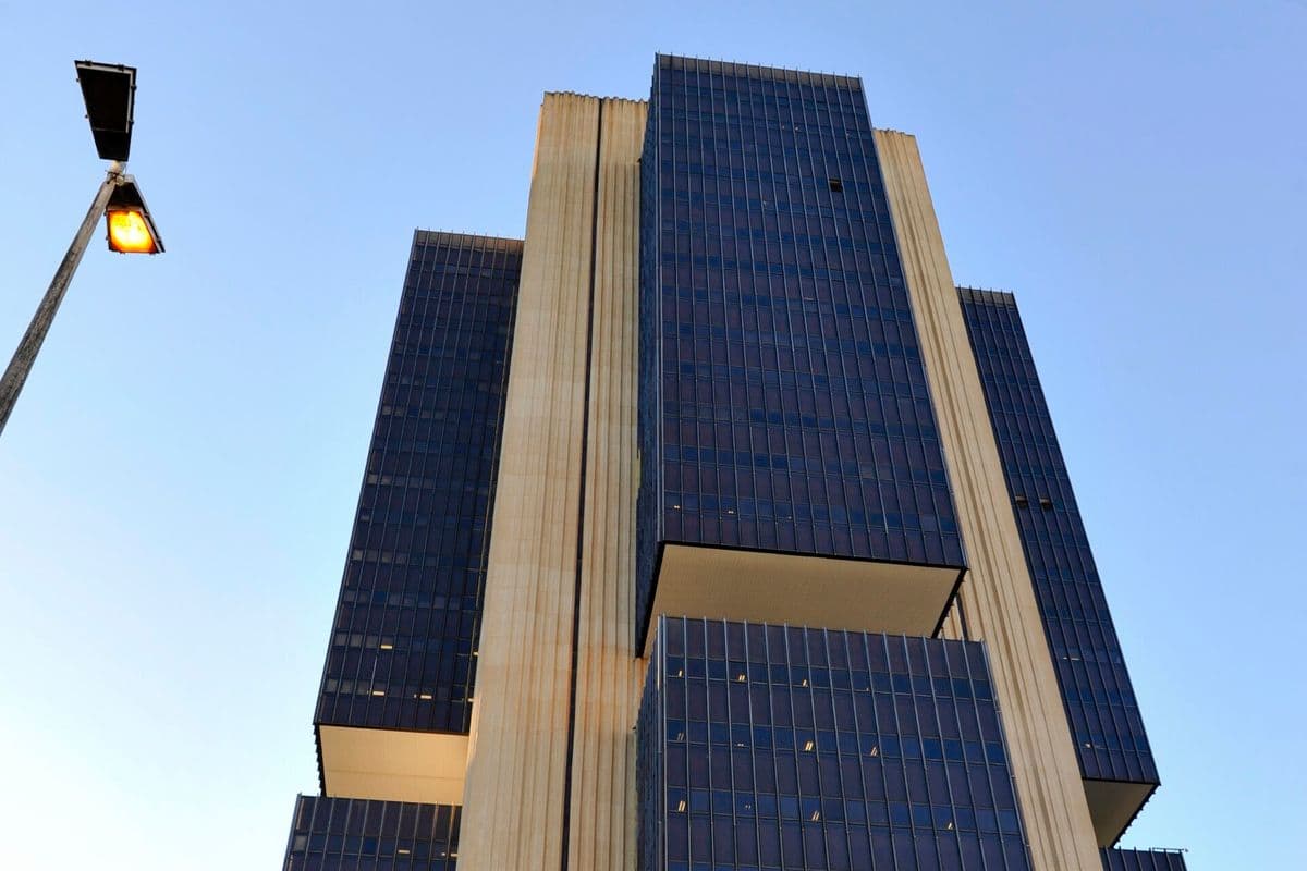 Edifício-sede do Banco Central visto de baixo para cima, com blocos de vidro azul-escuro e faixas verticais de concreto claro contra o céu azul, e um poste de luz aceso à esquerda