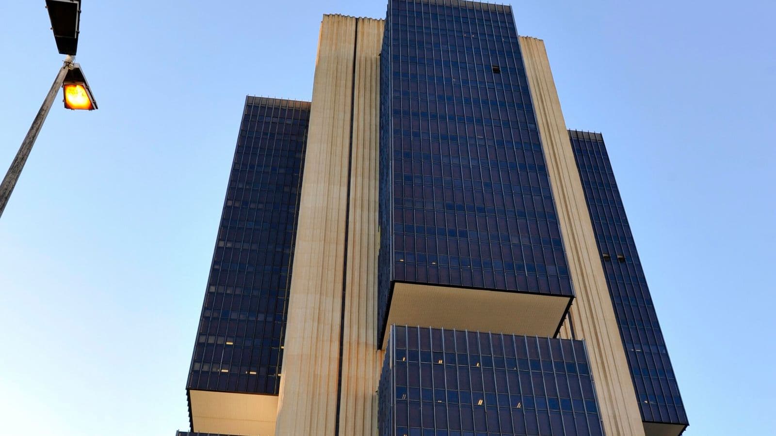 Edifício-sede do Banco Central visto de baixo para cima, com blocos de vidro azul-escuro e faixas verticais de concreto claro contra o céu azul, e um poste de luz aceso à esquerda