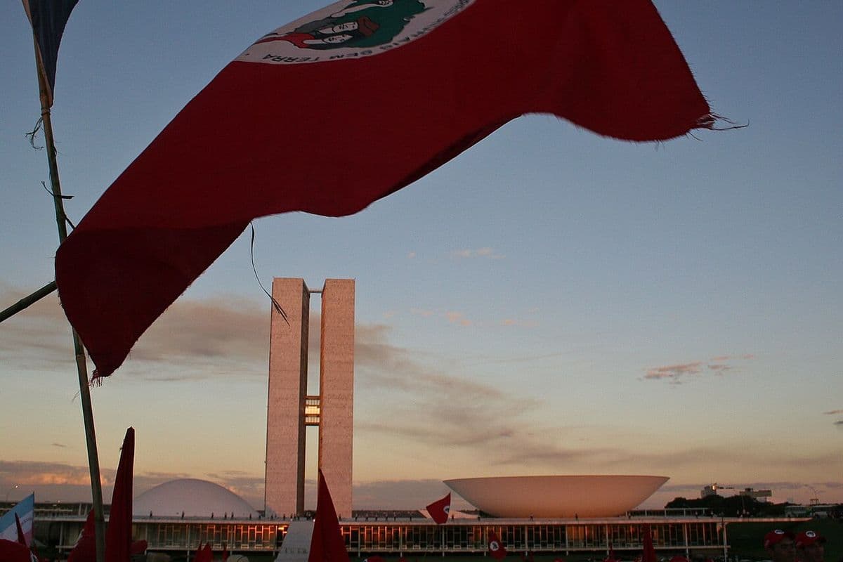 Bandeira vermelha do MST tremula em primeiro plano diante das torres e cúpulas do Congresso Nacional, com manifestantes de boné vermelho ao fundo