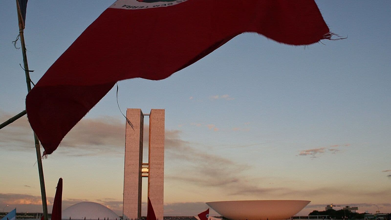 Bandeira vermelha do MST tremula em primeiro plano diante das torres e cúpulas do Congresso Nacional, com manifestantes de boné vermelho ao fundo