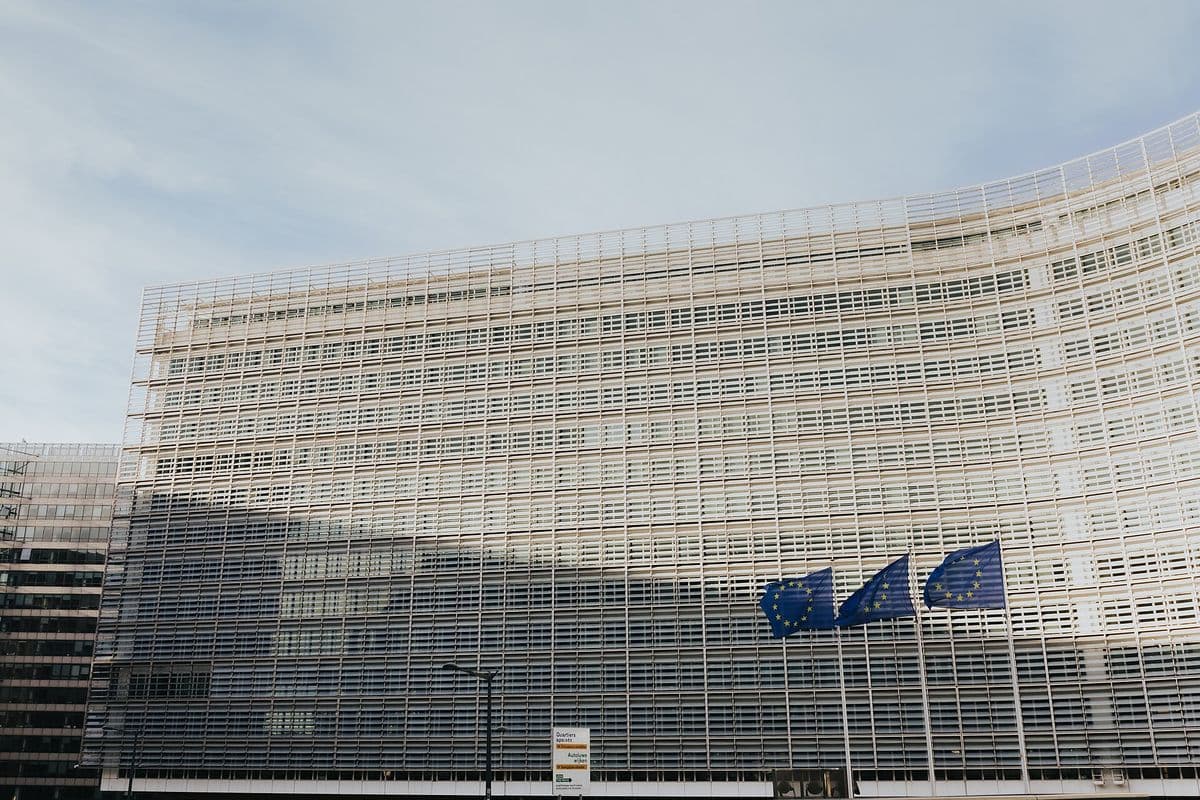 Fachada curva de vidro e aço do edifício Berlaymont, com três bandeiras azuis da União Europeia hasteadas à direita, sob céu claro