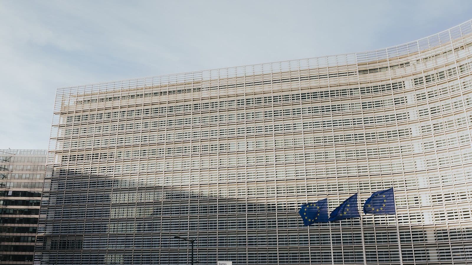 Fachada curva de vidro e aço do edifício Berlaymont, com três bandeiras azuis da União Europeia hasteadas à direita, sob céu claro
