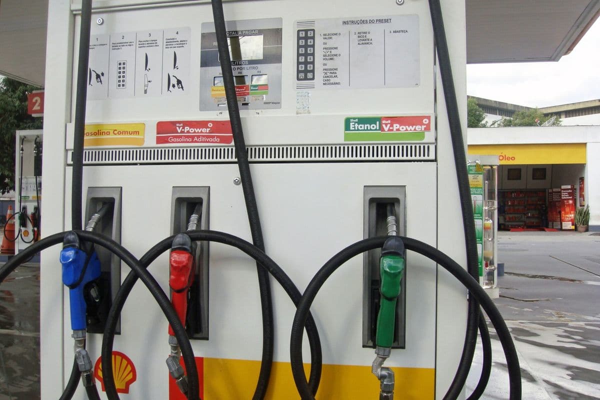 Bomba de combustível branca com três bicos: azul e vermelho para gasolina e verde para etanol, em posto coberto.