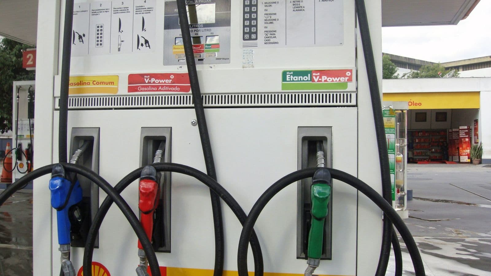 Bomba de combustível branca com três bicos: azul e vermelho para gasolina e verde para etanol, em posto coberto.