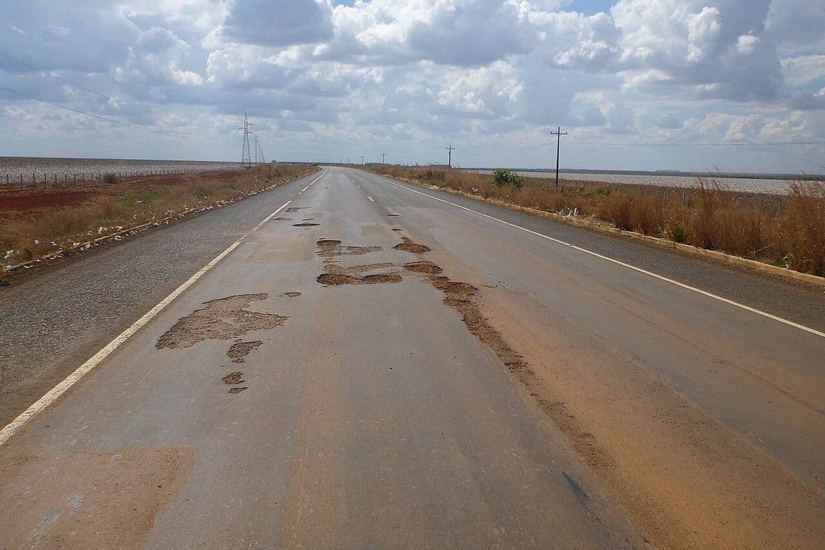 Rodovia de pista simples com remendos e buracos no asfalto, cortando lavouras planas sob céu com nuvens, com linhas de transmissão ao lado
