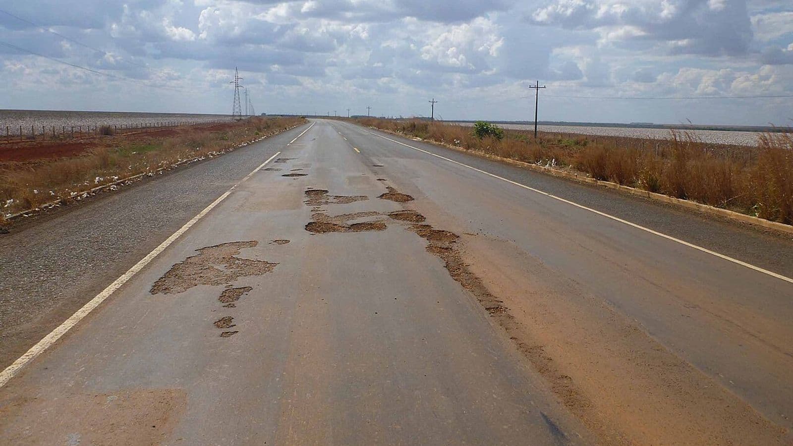 Rodovia de pista simples com remendos e buracos no asfalto, cortando lavouras planas sob céu com nuvens, com linhas de transmissão ao lado