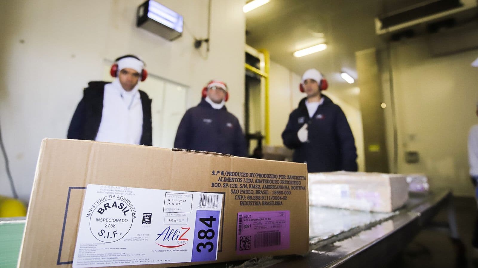 Caixa de papelão de frango congelado com selo do Serviço de Inspeção Federal do Ministério da Agricultura sobre uma esteira de frigorífico; ao fundo, três funcionários de uniforme térmico e protetor auricular.