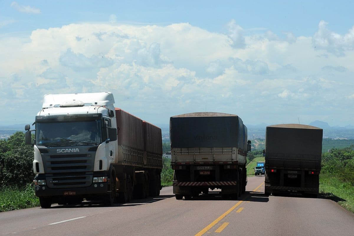 Três caminhões graneleiros com a carga coberta por lona trafegam por rodovia de pista simples, sob céu com nuvens