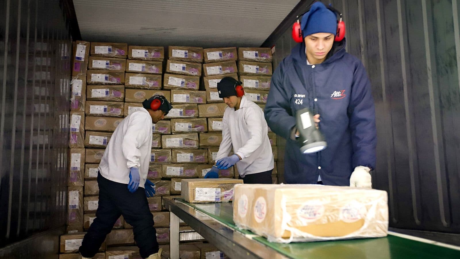 Dentro de um contêiner refrigerado forrado de caixas de papelão, dois funcionários de jaleco branco, luvas azuis e protetor auricular empilham caixas de frango congelado, enquanto um terceiro, de casaco azul e gorro, segura um leitor portátil junto a uma esteira com uma caixa.