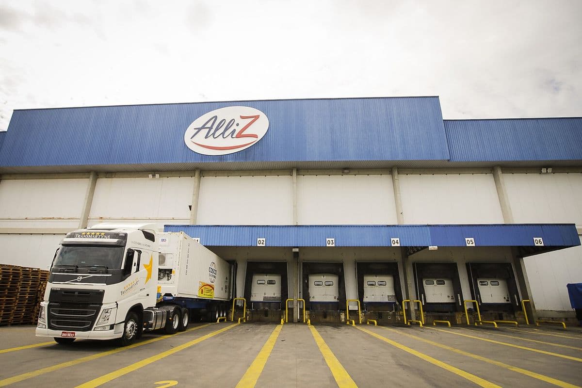 Caminhão branco com contêiner refrigerado encostado nas docas de um armazém frigorífico branco e azul, sob céu nublado