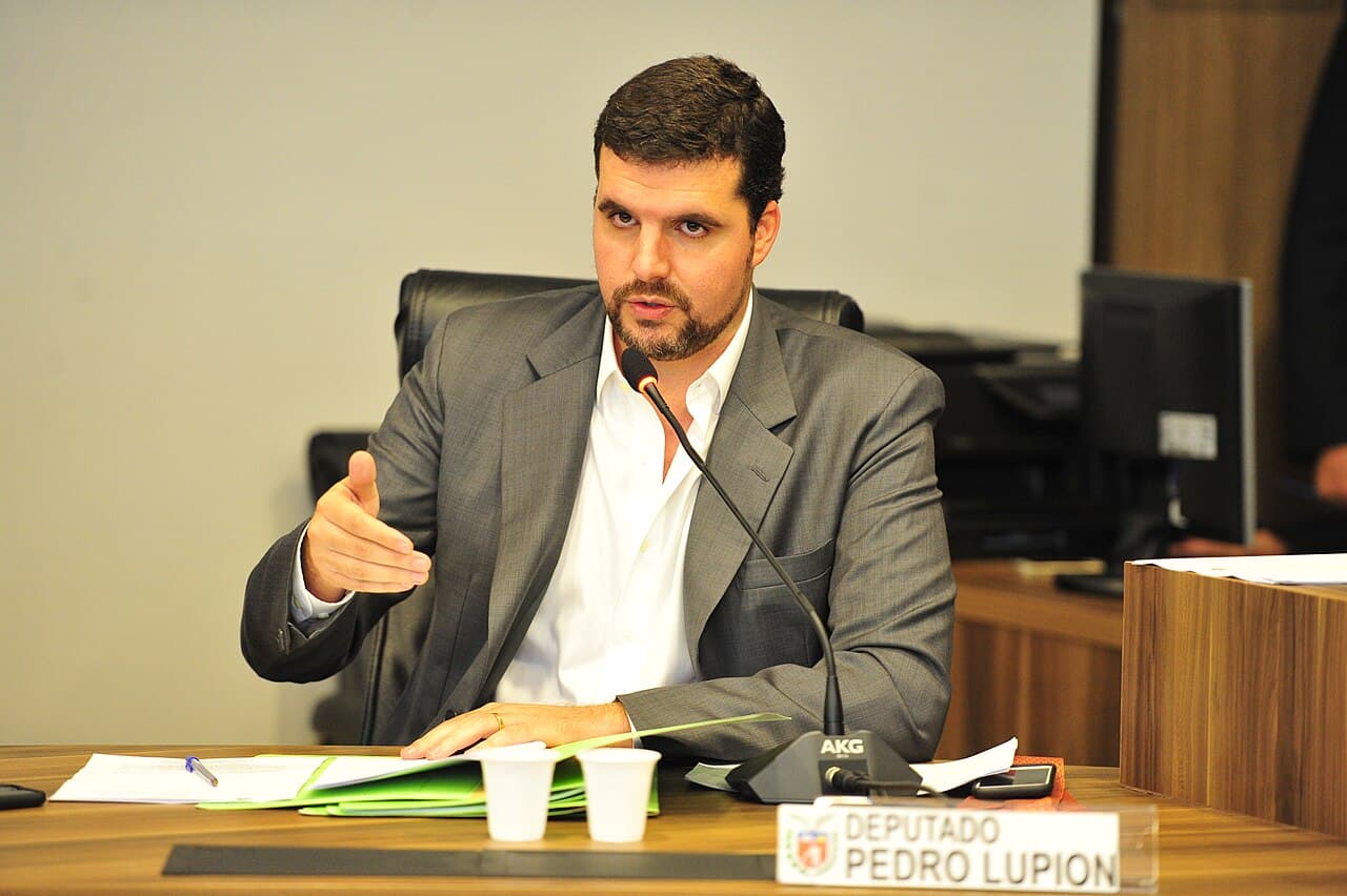 Pedro Lupion, de terno cinza e camisa branca, fala ao microfone sentado a uma mesa com a placa 'Deputado Pedro Lupion'