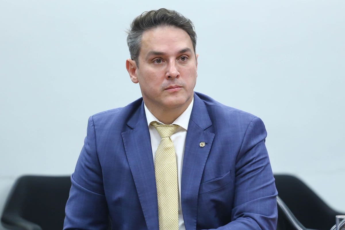 Homem de terno azul, camisa branca e gravata amarela, sentado diante de uma parede clara, com expressão séria.