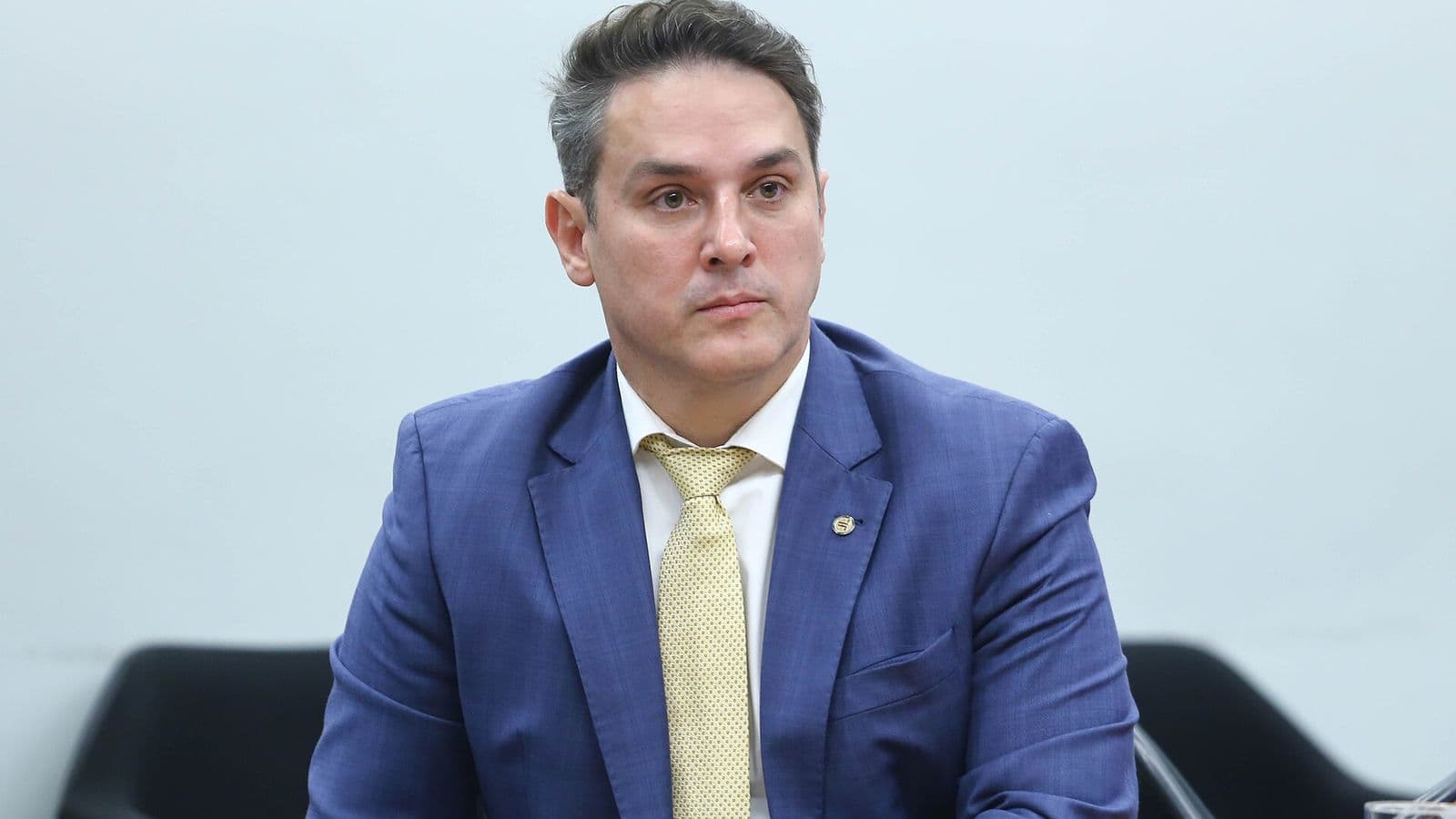 Homem de terno azul, camisa branca e gravata amarela, sentado diante de uma parede clara, com expressão séria.