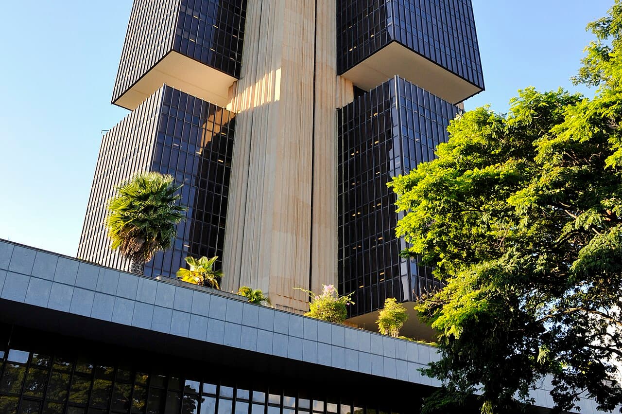 Torre de vidro escuro e concreto do edifício-sede do Banco Central, em Brasília, com palmeiras e uma árvore em primeiro plano
