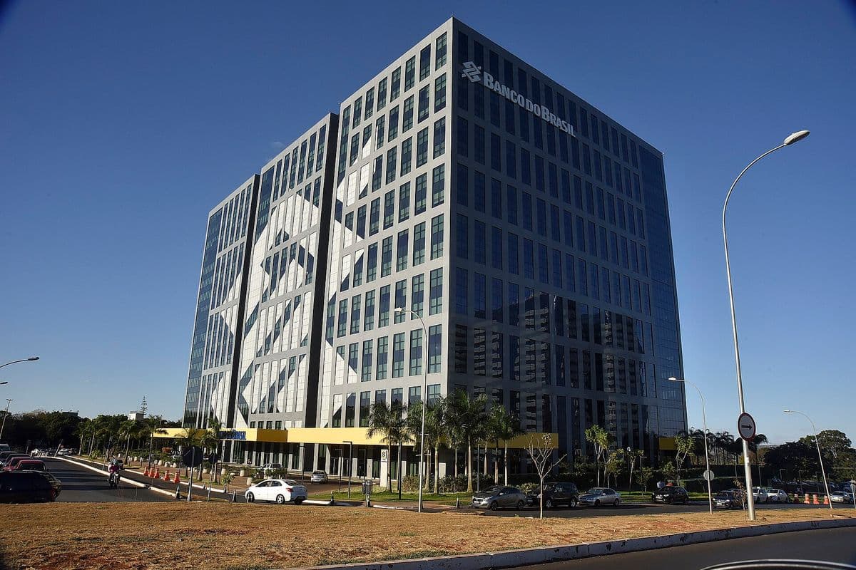 Prédio envidraçado de cerca de 15 andares com o logotipo do Banco do Brasil no alto da fachada, palmeiras e carros estacionados na frente, sob céu azul