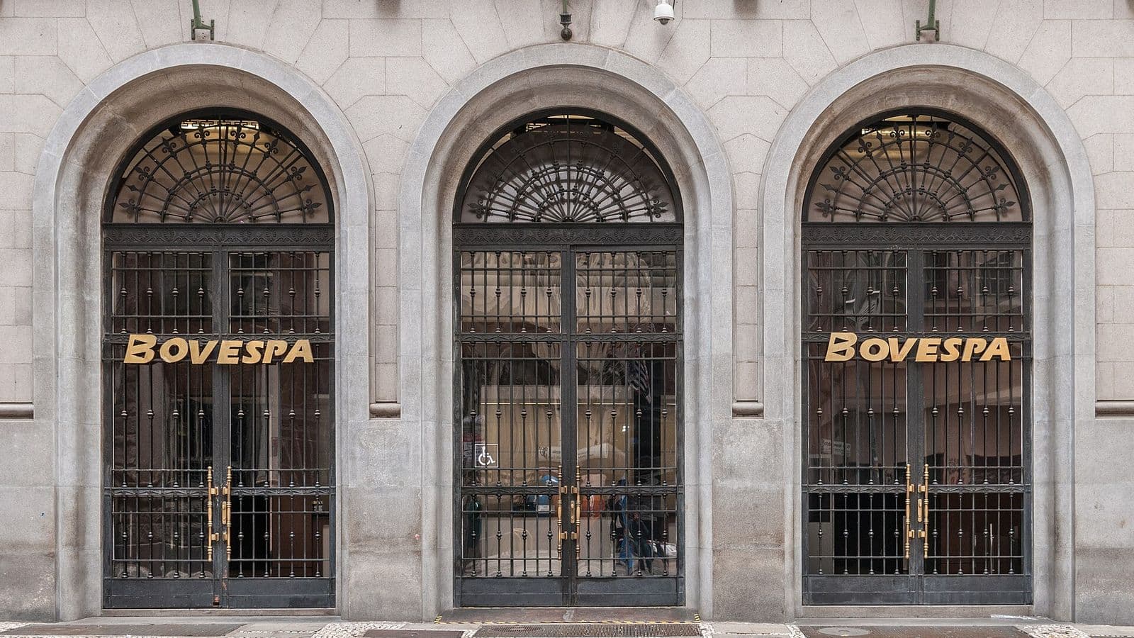 Fachada de pedra com três portões em arco de ferro trabalhado e o nome Bovespa em letras douradas sobre as grades, em rua de calçada de pedras portuguesas
