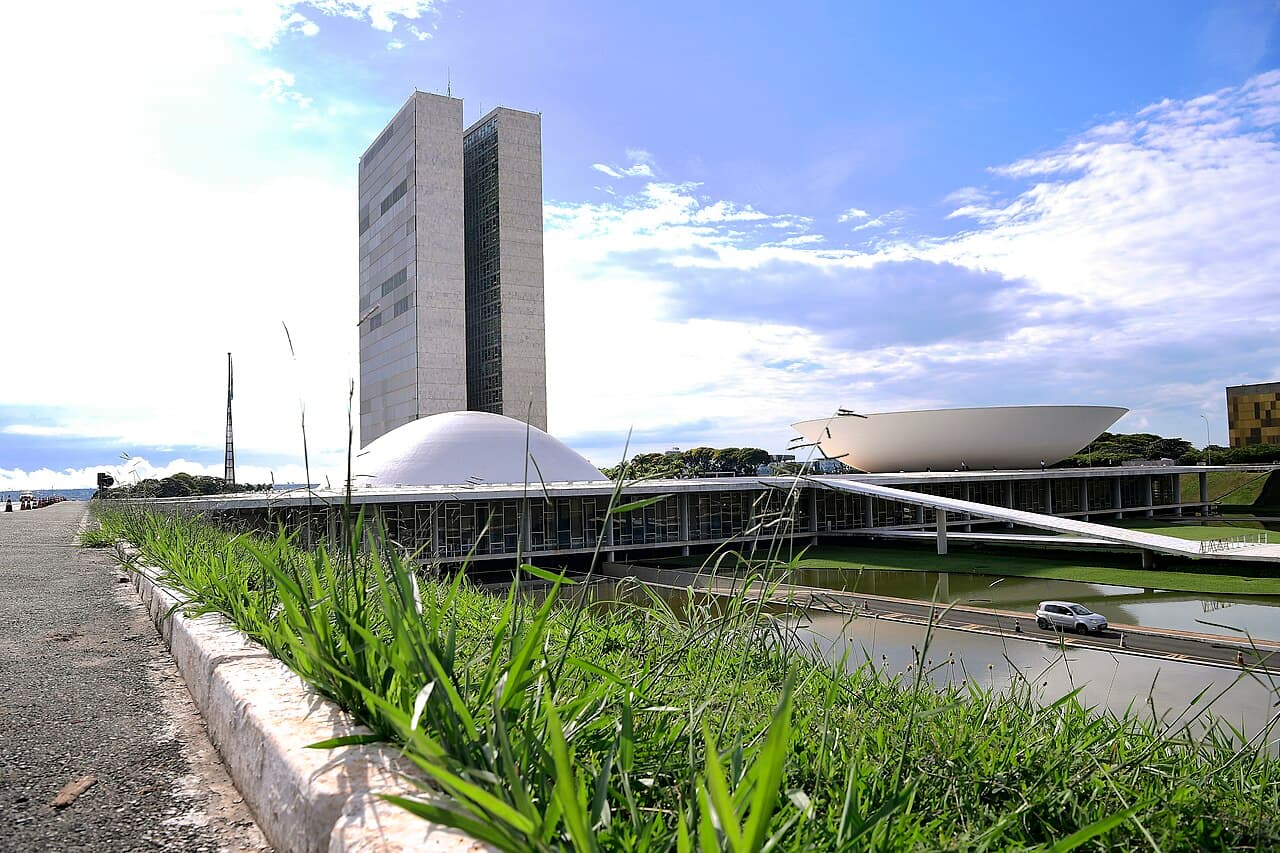 Torres do Congresso Nacional entre a cúpula do Senado e a cúpula invertida da Câmara, com o espelho d'água e gramado em primeiro plano