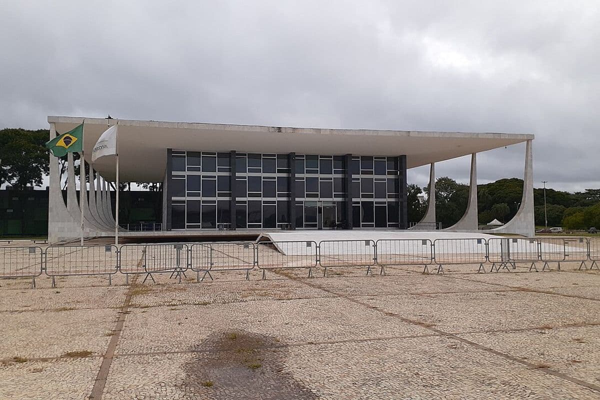 Fachada do Palácio do Supremo Tribunal Federal, com colunas curvas brancas, bandeira do Brasil e grades na praça em frente, sob céu nublado
