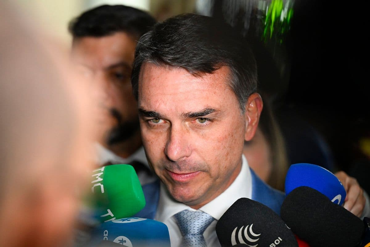 Flávio Bolsonaro, de terno azul, camisa branca e gravata cinza, fala de frente para a câmera cercado de microfones de emissoras de TV.