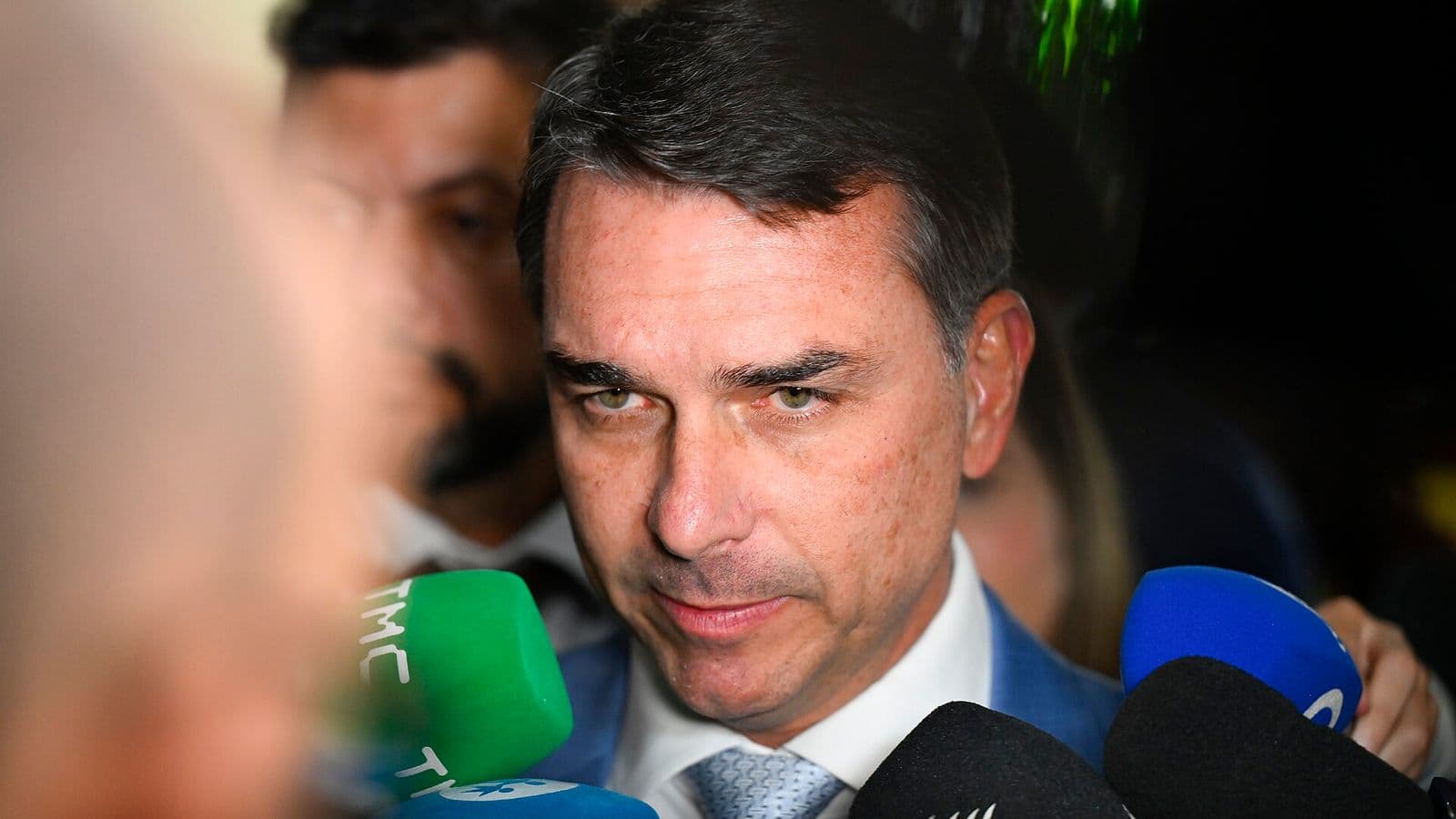 Flávio Bolsonaro, de terno azul, camisa branca e gravata cinza, fala de frente para a câmera cercado de microfones de emissoras de TV.