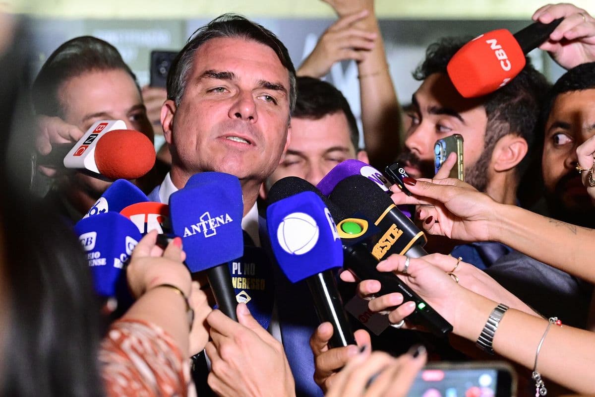 Flávio Bolsonaro, de terno escuro, fala cercado de microfones de várias emissoras e de celulares estendidos por repórteres.