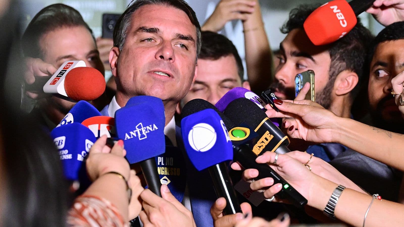 Flávio Bolsonaro, de terno escuro, fala cercado de microfones de várias emissoras e de celulares estendidos por repórteres.