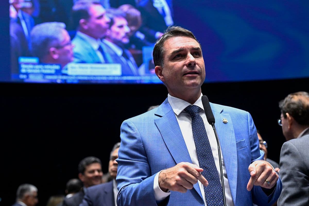 Flávio Bolsonaro, de terno azul-claro e gravata azul com bolinhas, fala ao microfone de pé no plenário do Senado, com um telão ao fundo.