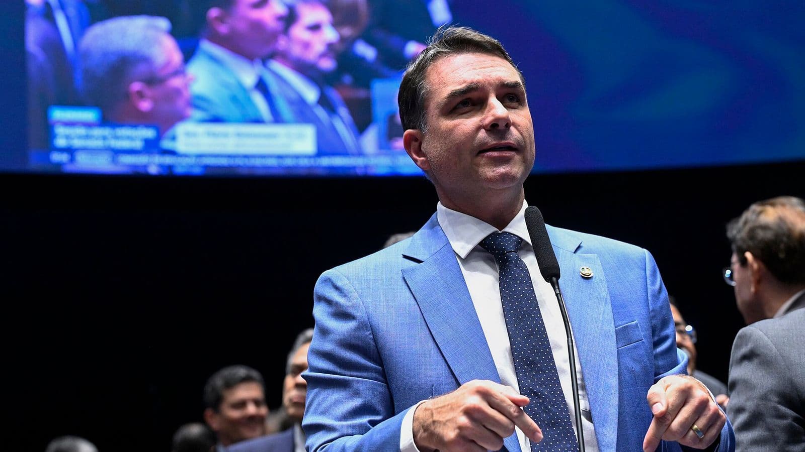 Flávio Bolsonaro, de terno azul-claro e gravata azul com bolinhas, fala ao microfone de pé no plenário do Senado, com um telão ao fundo.