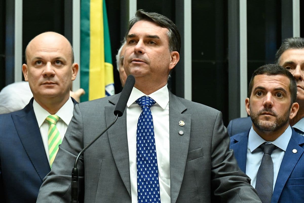 Flávio Bolsonaro, de terno cinza e gravata azul, fala ao microfone na tribuna do plenário, com parlamentares de terno atrás dele e uma bandeira do Brasil ao fundo.