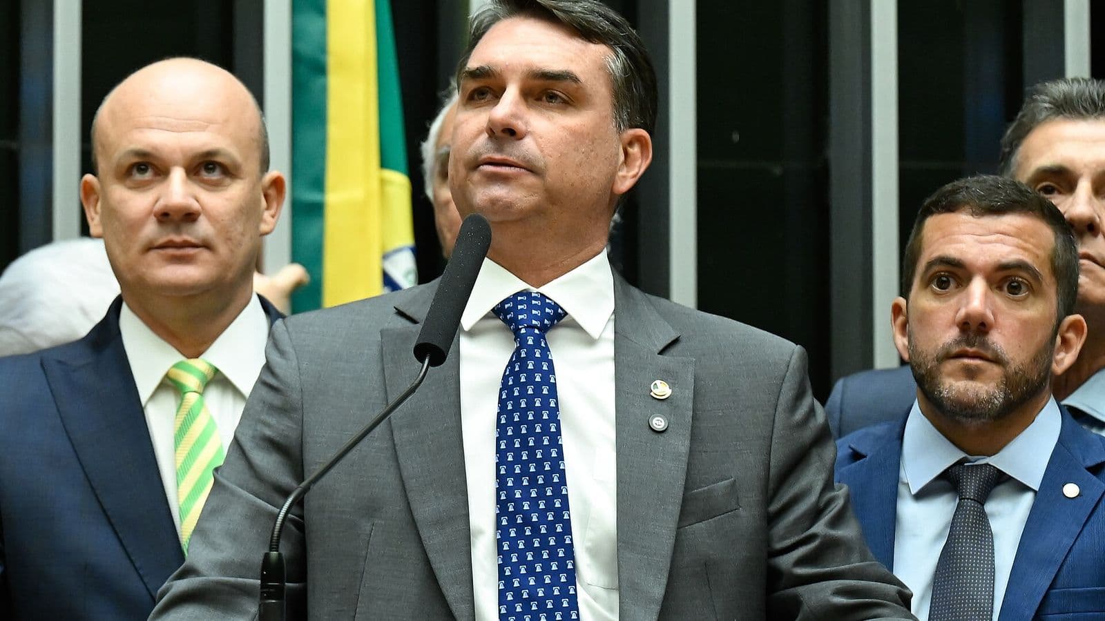 Flávio Bolsonaro, de terno cinza e gravata azul, fala ao microfone na tribuna do plenário, com parlamentares de terno atrás dele e uma bandeira do Brasil ao fundo.