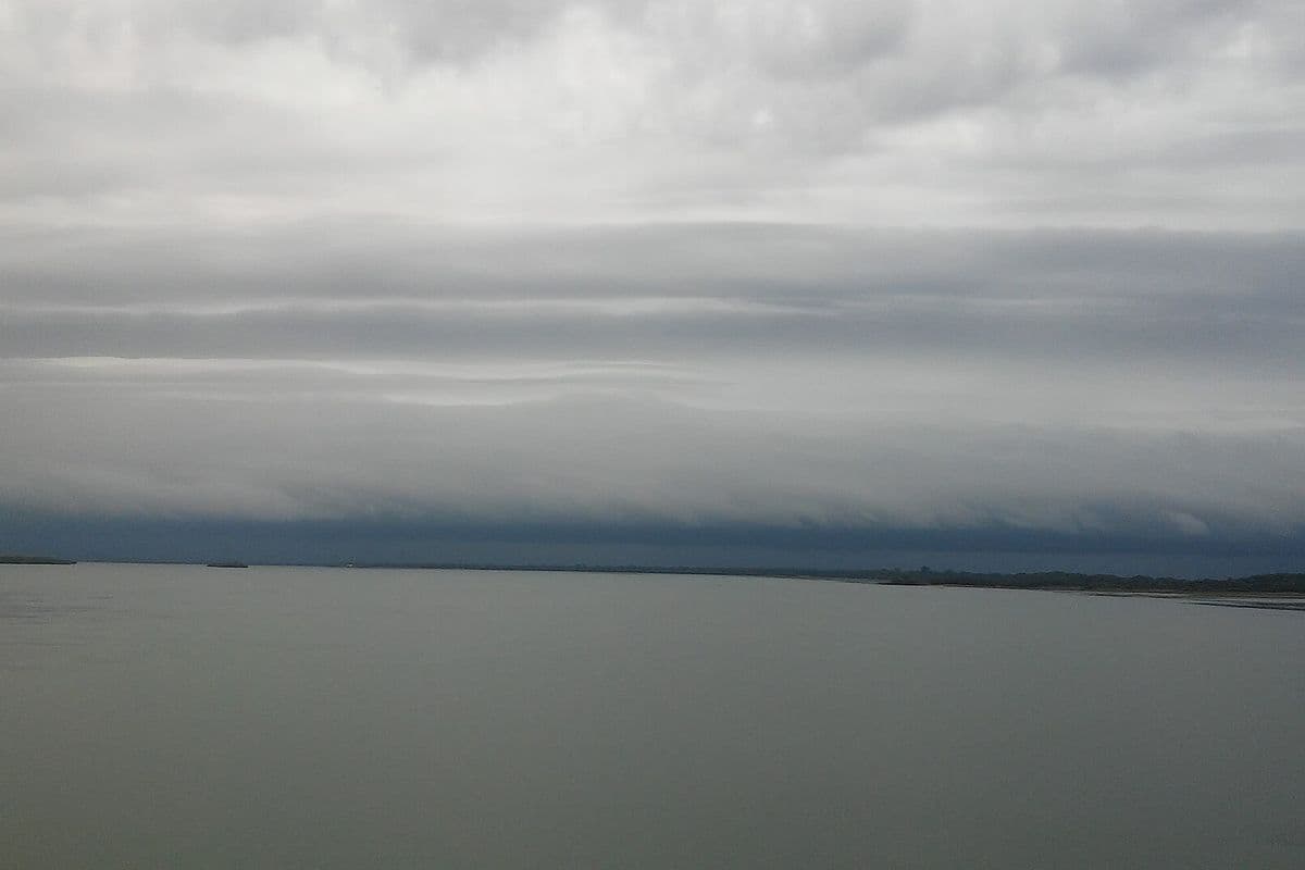 Faixa espessa de nuvens cinza-escuras, em camadas horizontais, avança no horizonte sobre a superfície larga e calma de um rio, sob céu encoberto