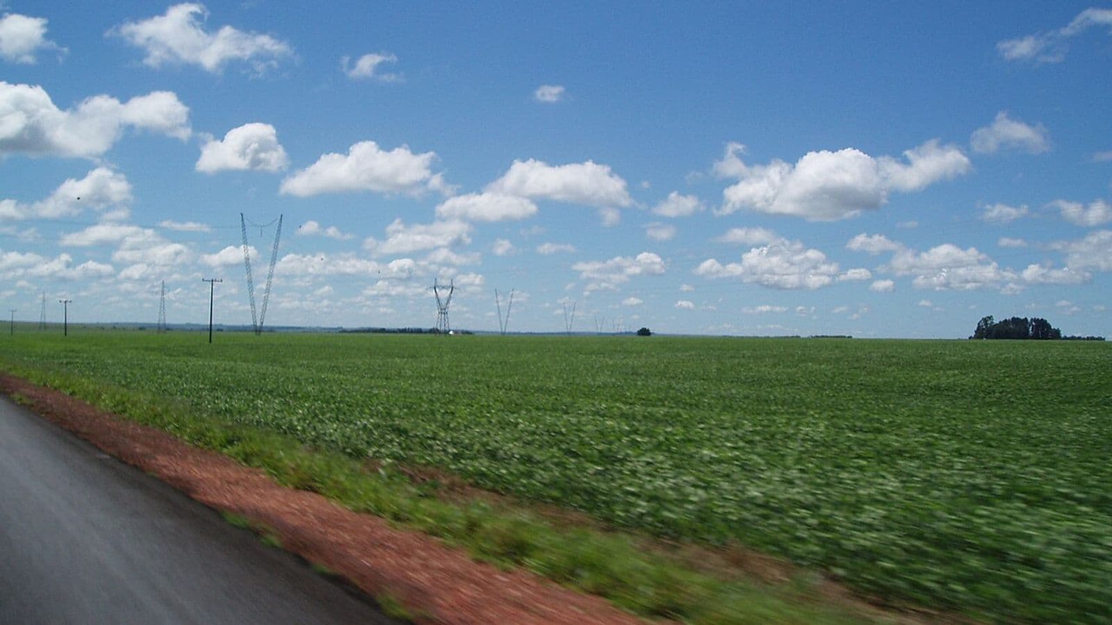 Lavoura de soja verde e plana à beira de uma estrada asfaltada com acostamento de terra vermelha, sob céu azul com nuvens e torres de linha de transmissão ao fundo