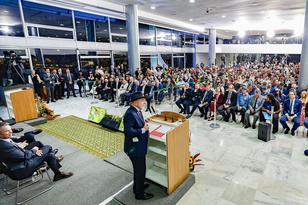 Lula, de terno escuro e chapéu, discursa em um púlpito de madeira diante de uma plateia sentada em um salão amplo com piso de mármore e paredes de vidro.
