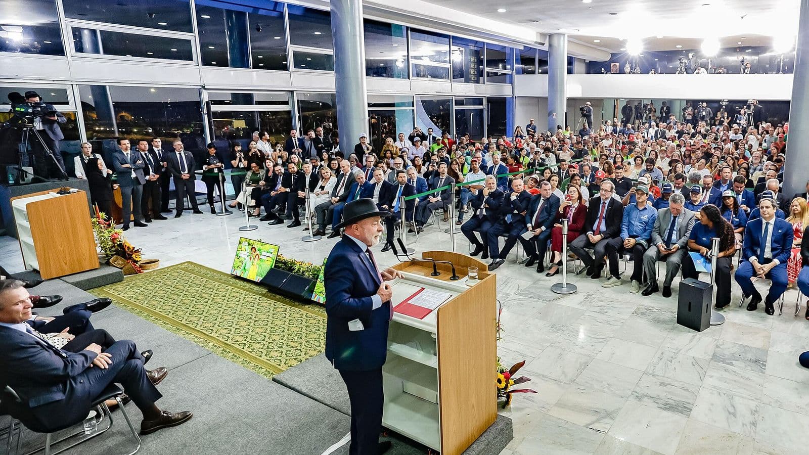 Lula, de terno escuro e chapéu, discursa em um púlpito de madeira diante de uma plateia sentada em um salão amplo com piso de mármore e paredes de vidro.