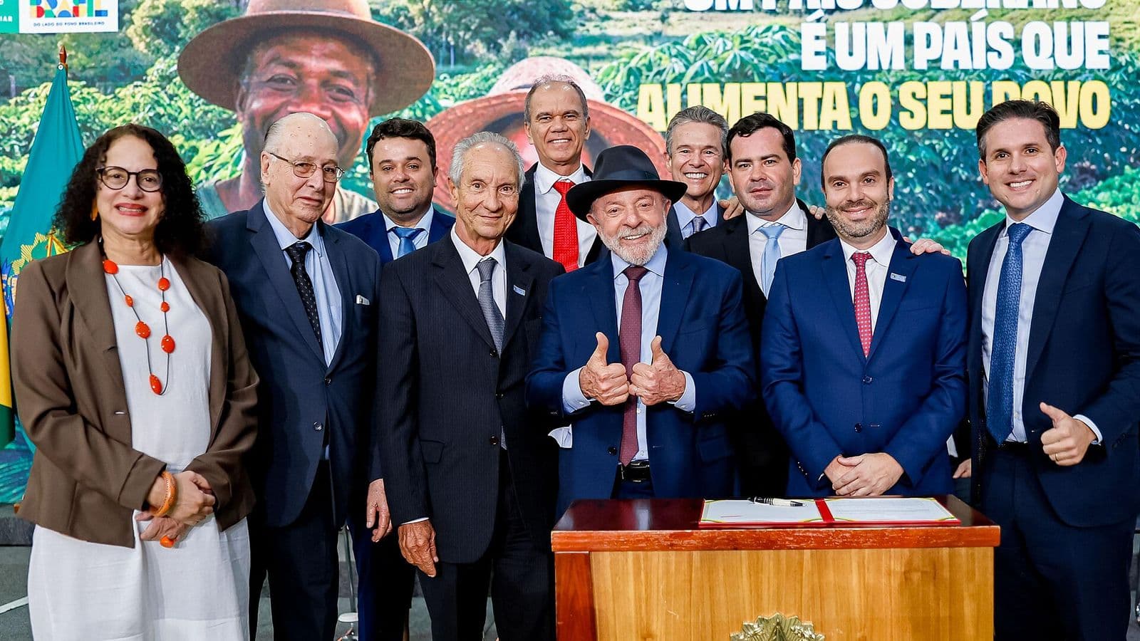 Lula, de chapéu preto e terno azul, faz sinal de positivo com as duas mãos ao lado de ministros e autoridades, diante de um púlpito com o brasão da República e de um painel com a frase "Um país soberano é um país que alimenta o seu povo".
