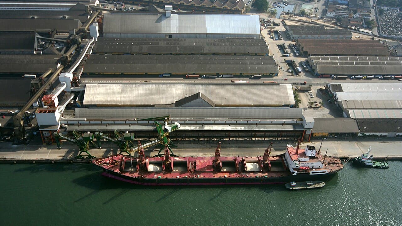 Vista aérea de um navio graneleiro com porões abertos atracado diante de armazéns e esteiras de um terminal de grãos