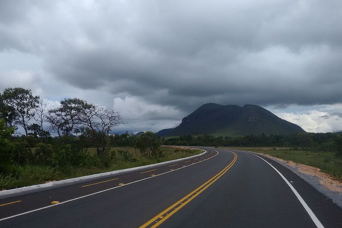 Rodovia asfaltada faz curva em direção a um morro, sob céu fechado por nuvens escuras de chuva, com vegetação de cerrado nas margens