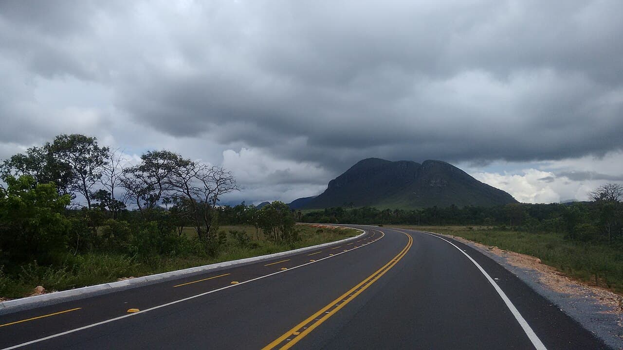 Rodovia asfaltada faz curva em direção a um morro, sob céu fechado por nuvens escuras de chuva, com vegetação de cerrado nas margens