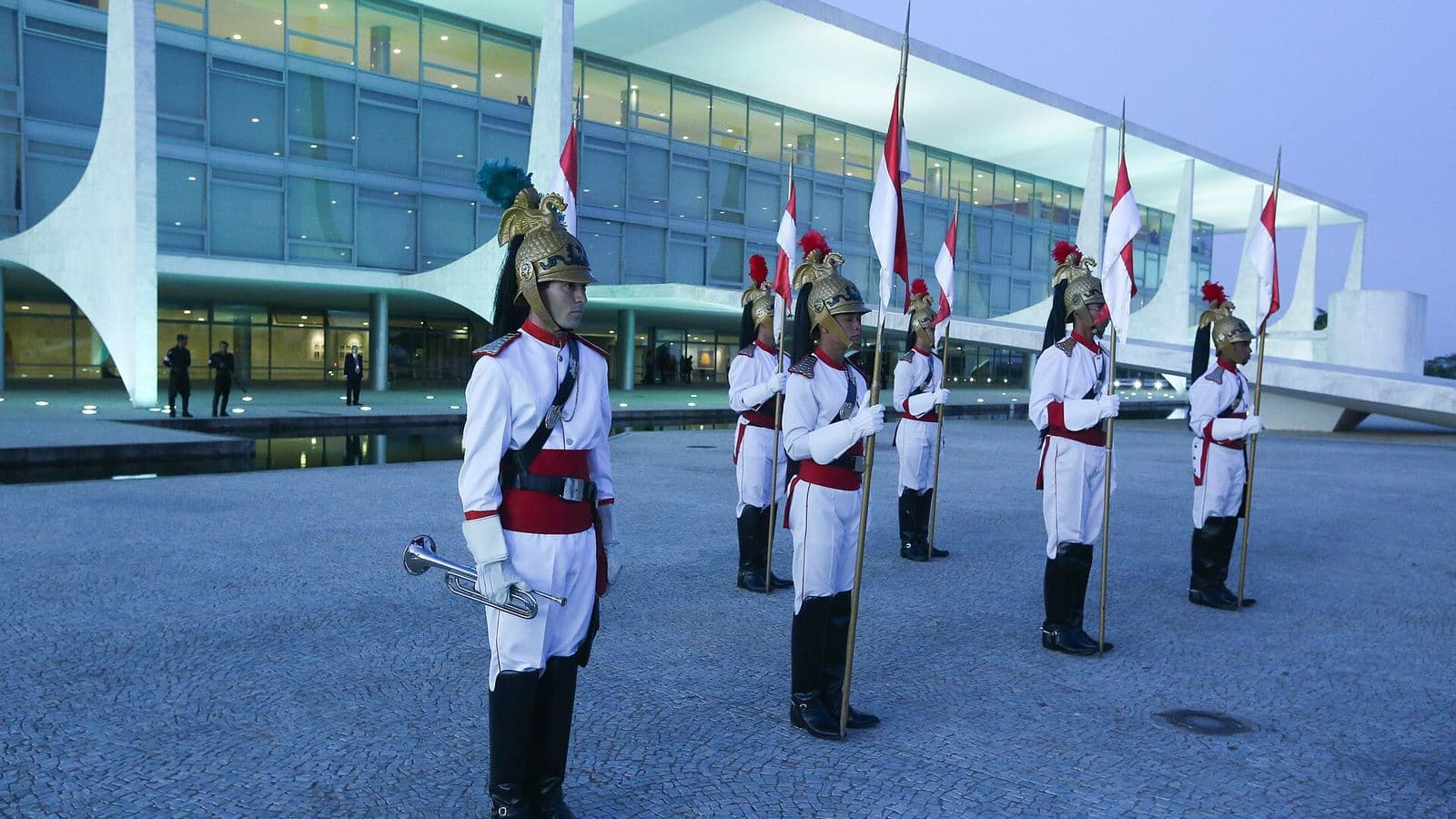 Cinco soldados dos Dragões da Independência, de uniforme branco e vermelho e capacete dourado com penacho, perfilados com lanças diante da fachada iluminada do Palácio do Planalto ao anoitecer