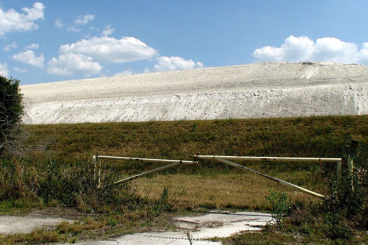 Grande morro esbranquiçado de fosfogesso ao fundo, atrás de um campo de grama e de uma porteira de metal, sob céu azul com nuvens