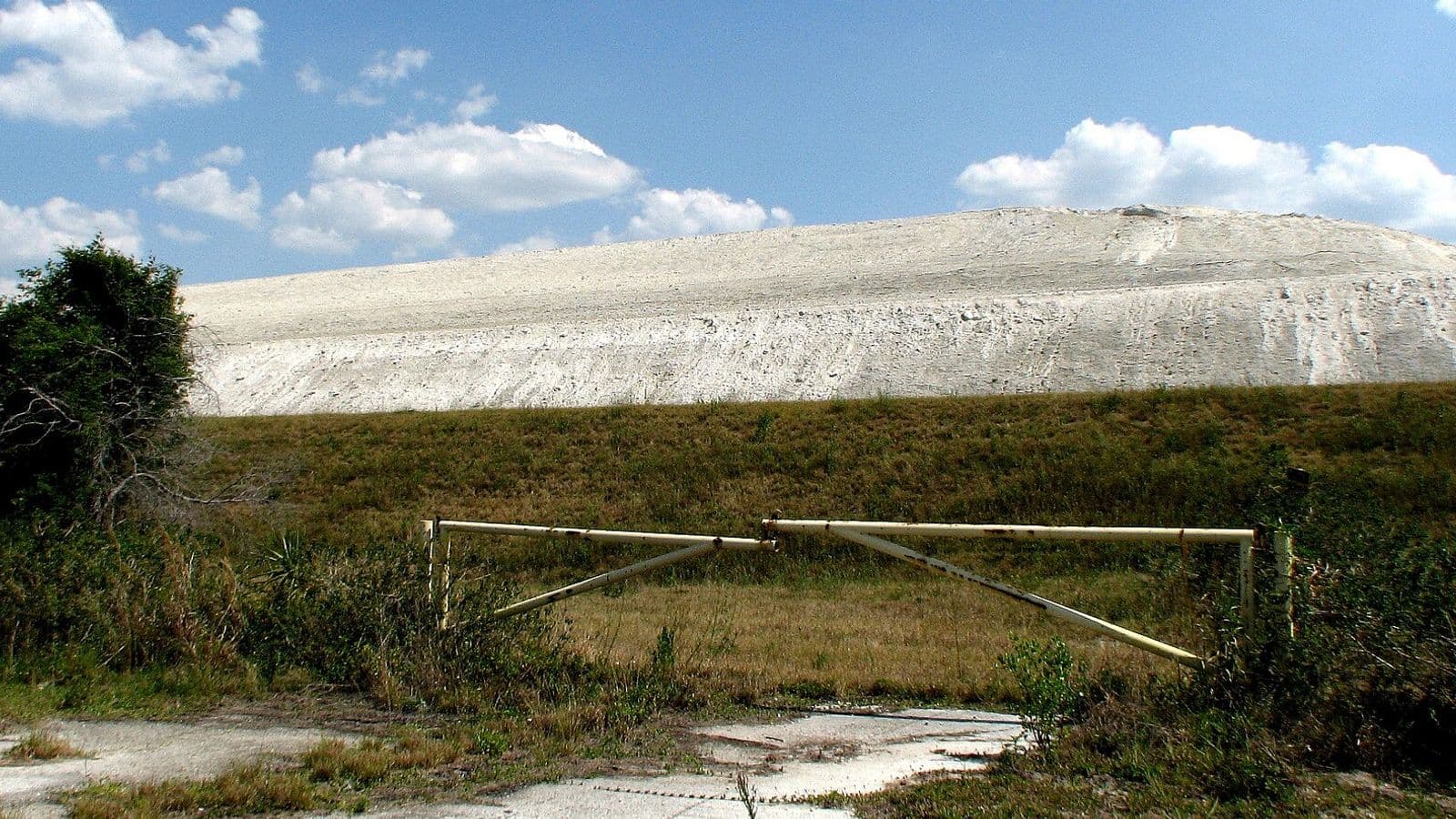 Grande morro esbranquiçado de fosfogesso ao fundo, atrás de um campo de grama e de uma porteira de metal, sob céu azul com nuvens