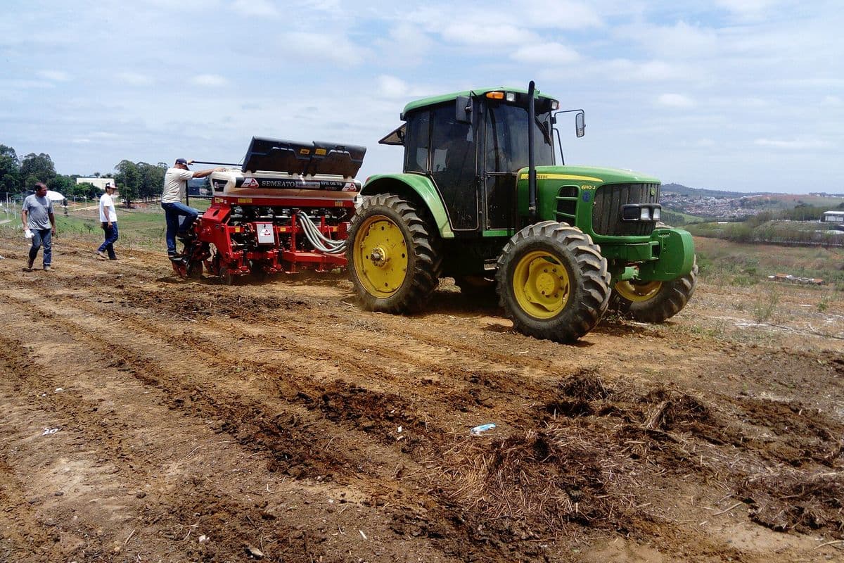 Trator verde puxa uma plantadeira vermelha sobre terra revolvida, enquanto três homens acompanham o plantio
