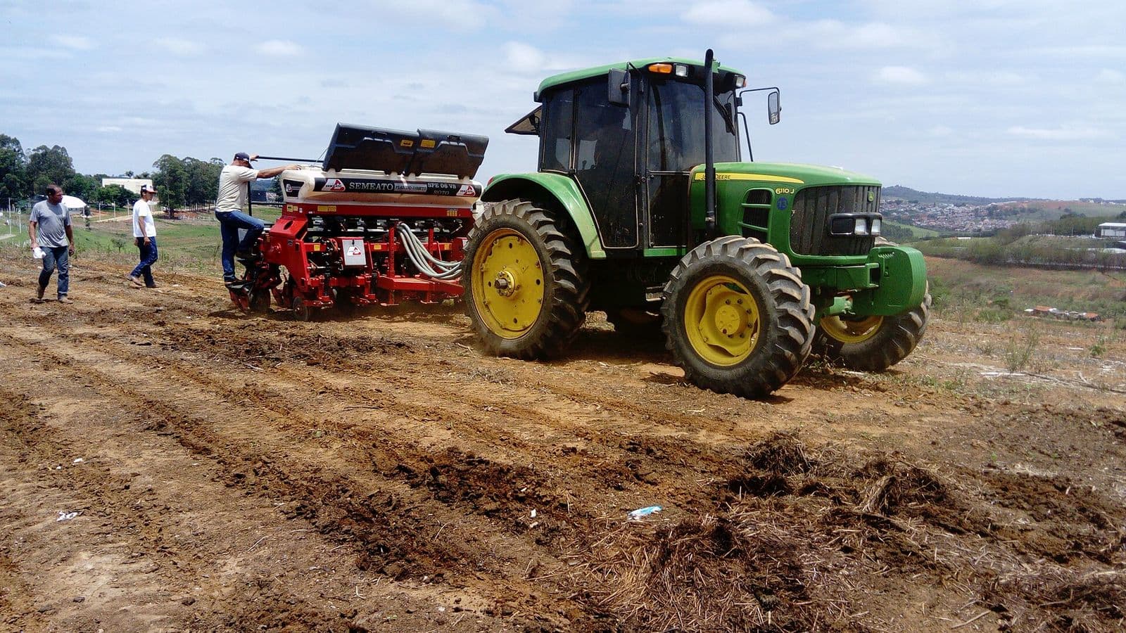Trator verde puxa uma plantadeira vermelha sobre terra revolvida, enquanto três homens acompanham o plantio