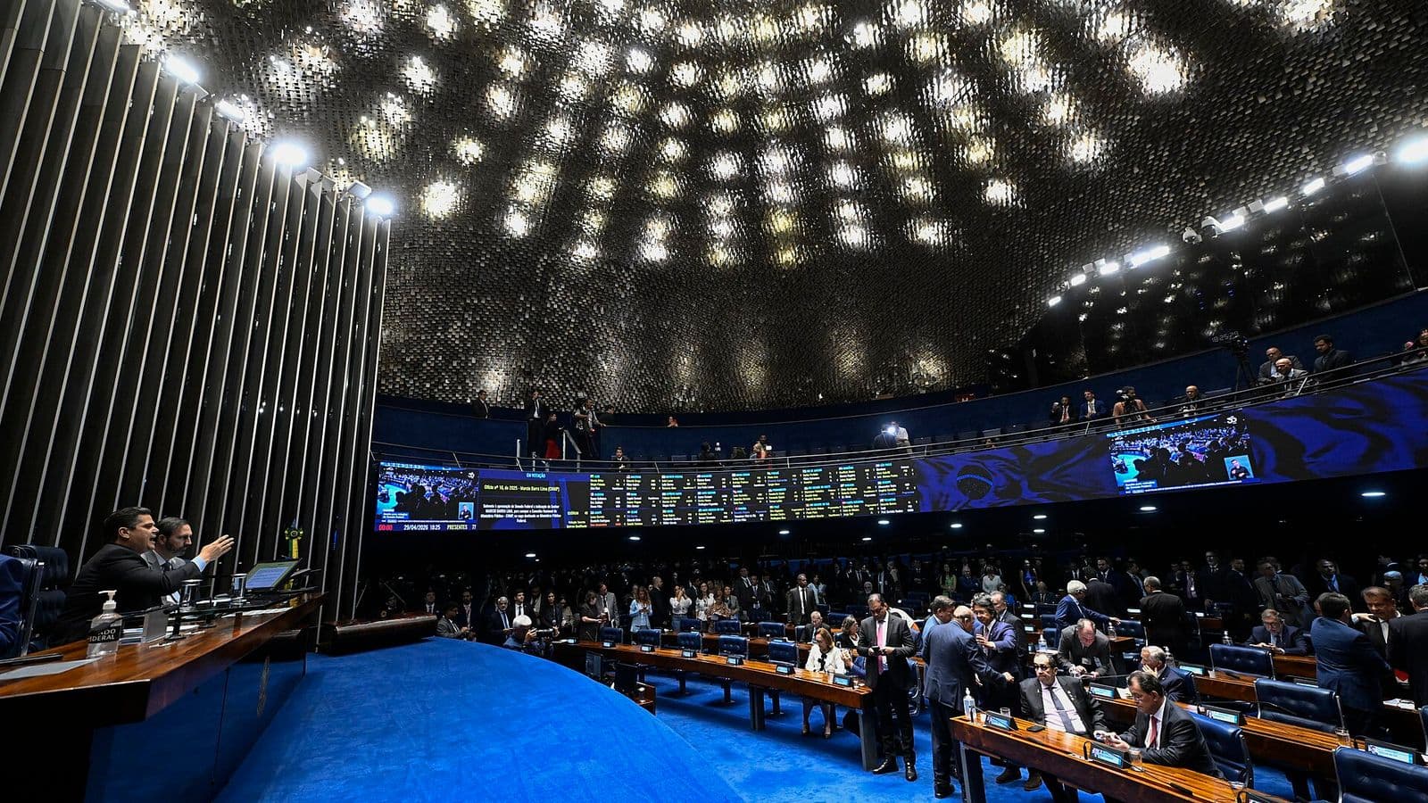Plenário do Senado visto do alto, com senadores em pé entre as bancadas azuis e o painel eletrônico de votação ao fundo