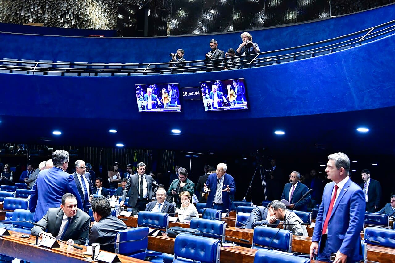 Plenário do Senado com senadores nas bancadas de cadeiras azuis, alguns de pé conversando, e fotógrafos na galeria