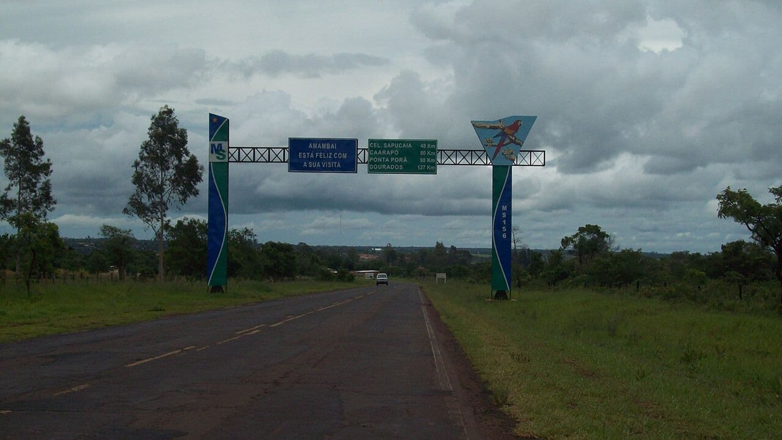 Rodovia de asfalto passa sob pórtico azul e verde com placas de boas-vindas e de distâncias, sob céu carregado de nuvens